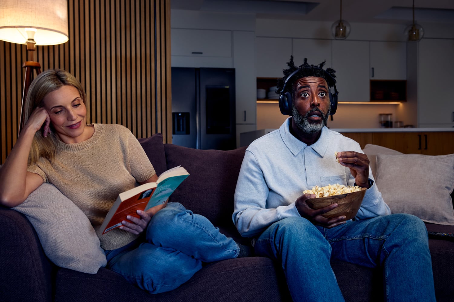 Zwei Personen auf einem Sofa, die rechte schaut TV mit Kopfhörer, die linke liest